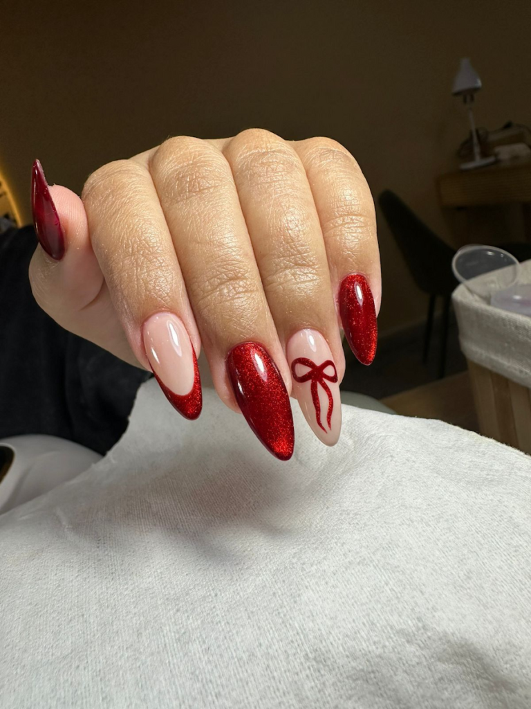 Primer plano de una mano con una manicura francesa clásica en forma de almendra, donde las puntas son de un rojo brillante con purpurina y una uña presenta un delicado lazo rojo pintado.