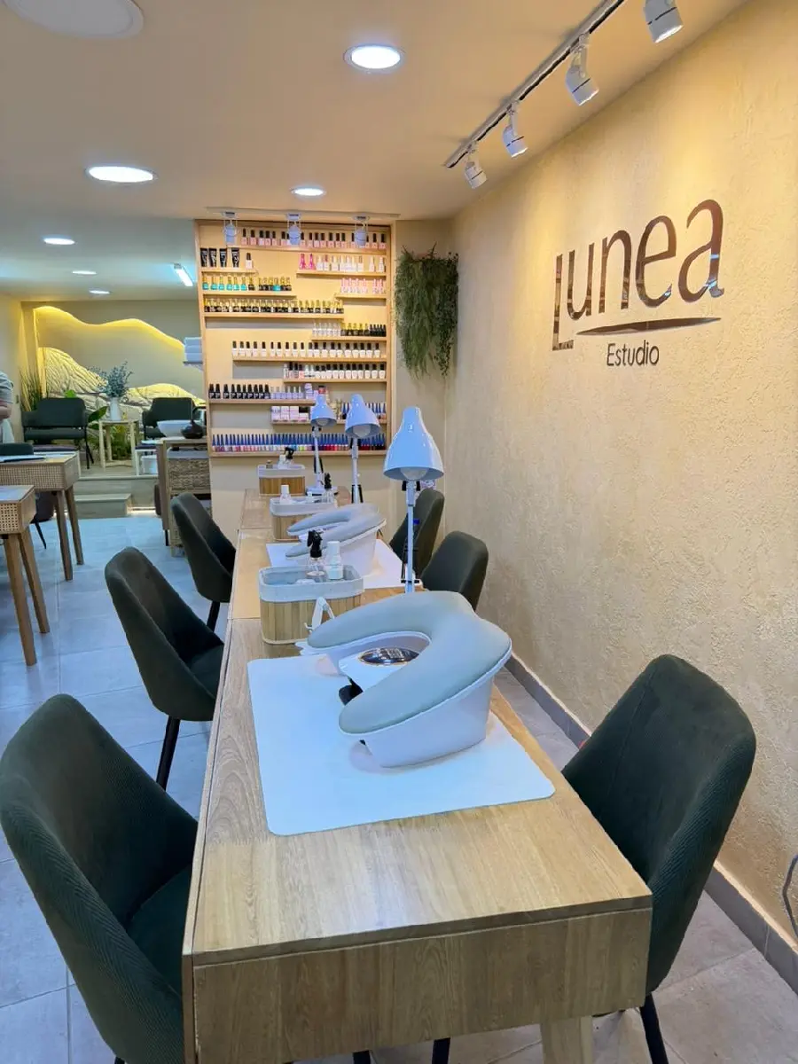 Vista general del salón de manicura Lunea Estudio, mostrando tanto la zona de manicura con mesas y sillas como la zona de pedicura elevada al fondo, con la pared de piedra retroiluminada y las plantas verdes
